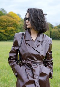 Barna, fényes, övvel ellátott esernyőkabát széles hajtókákkal és oldalzsebekkel. A modellen kis fekete napszemüveg van, és egy fekete táskát tart arany kiegészítőkkel.