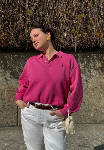 Mujer con cabello oscuro, camisa rosa de manga larga y pantalones blancos, de pie al aire libre contra una pared de hormigón con enredaderas sin hojas.