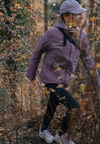 Giacca impermeabile viola con cerniera frontale, dettagli neri e logo. Indossata con leggings neri e scarpe da ginnastica chiare tra il fogliame autunnale.