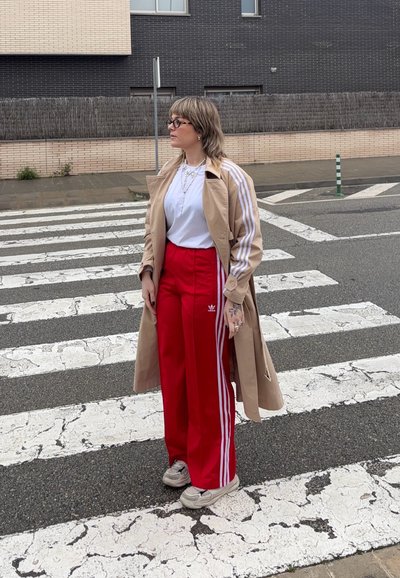 Abrigo trench beige con rayas blancas, camiseta blanca y pantalones de chándal rojos con rayas laterales blancas. Zapatillas grises, de pie en un paso de peatones.
