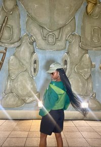Felpa verde e nera con un design a blocchi di colore, abbinata a pantaloni corti neri. Murale privo di caratteristiche sullo sfondo, illuminato da luci soffuse.