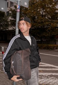 Jauns vīrietis melnā Adidas jaku un cepuri tur lielu maisu, stāvot pie ceļazīmes pie g�ājēju pārejas pilsētas ielā.