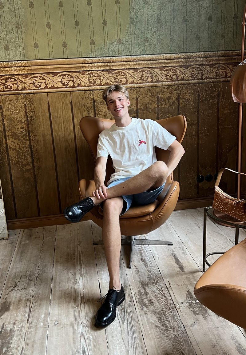 Jeune homme en t-shirt blanc et short assis en tailleur sur un fauteuil marron dans une pièce de style vintage avec un sol en bois et des panneaux muraux décoratifs.