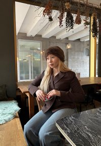 Nainen, jolla on ruskea viiruinen bleiseri ja turkislakki, istuu puupöydän ääressä pitäen kuppia käsissä�än. Hänellä on pitkät vaaleat hiukset ja päällään rentoja farkkuja.