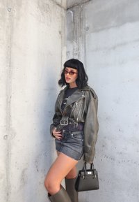 Zielona skórzana kurtka motocyklowa, czarny t-shirt z grafiką, postrzępione dżinsowe szorty, szare botki do kolan i mała czarna torebka. Brązowe okulary przeciwsłoneczne.