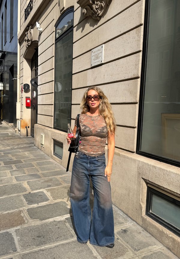 Mujer con cabello rubio largo que lleva gafas de sol, top transparente, jeans de pierna ancha, sosteniendo una botella de refresco de vidrio en una calle soleada de la ciudad.