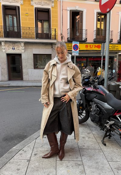 Gabardina beige sobre una sudadera con capucha gris, combinada con culottes de mezclilla oscura y botas marrones hasta la rodilla. Anillos metálicos adornan los dedos.