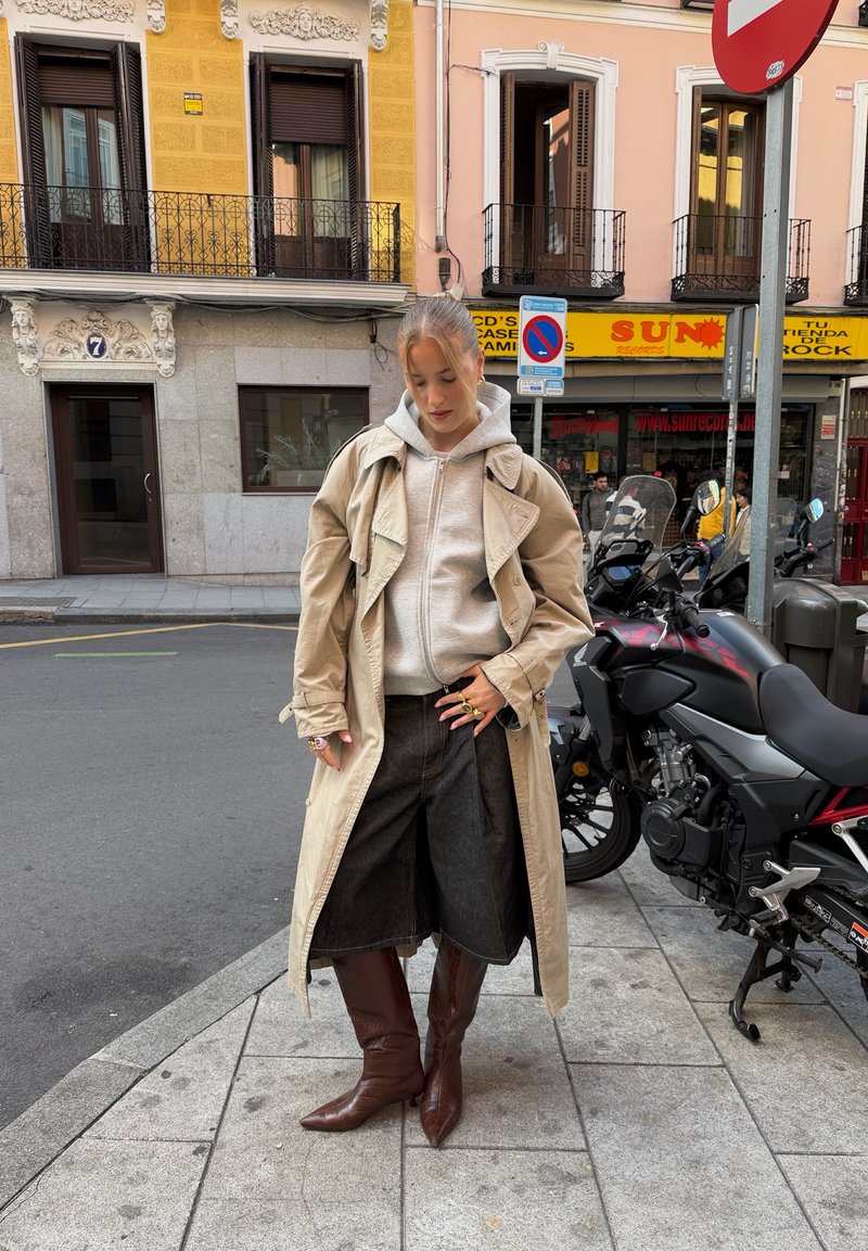 Trench-coat beige par-dessus un sweat à capuche gris, associé à des culottes en denim foncé et des bottes marron montant jusqu'aux genoux. Des bagues métalliques ornent les doigts.