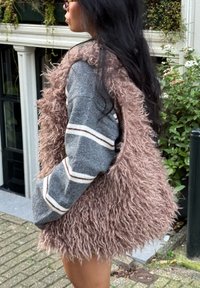 Pluizig roze gilet met een geschubde textuur over een grijze trui met witte en bruine strepen. De achtergrond toont groen en een gebouw.