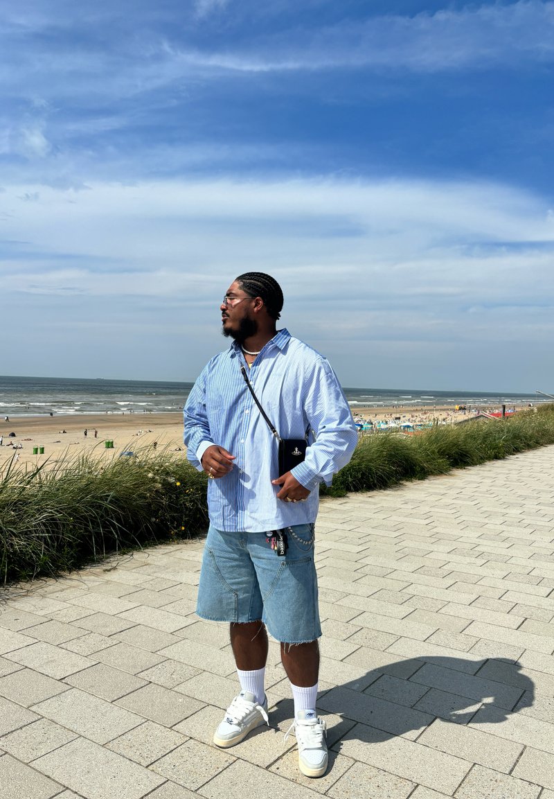 Μπλε και λευκή ριγέ πουκάμισο με κουμπιά, τζιν σορτς, λευκά αθλητικά παπούτσια και μια μικρή μαύρη τσάντα φορεμένη διαγωνίως μπροστά σε σκηνικό παραλίας.