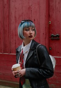 Manteau en cuir noir, chemise blanche à col, cravate à carreaux rouges, tenant une tasse de café. Lunettes de soleil rondes et sac en chaîne argenté. Coiffure bob bleue.