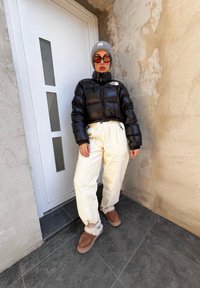 Μαύρο φούτερ μπουφάν, άσπρο παντελόνι με συγκεντρωμένα ποδόγυρα, καφέ αφράτες παντόφλες, γκρι καπέλο, και γυαλιά ηλίου. Στέκεται μπροστά από μια λευκή πόρτα.