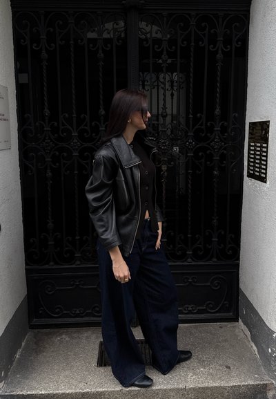 Chaqueta de cuero negra con un collar ancho, combinada con una blusa negra ajustada y pantalones de mezclilla oscuros anchos. Accesorios de pequeños pendientes de oro.