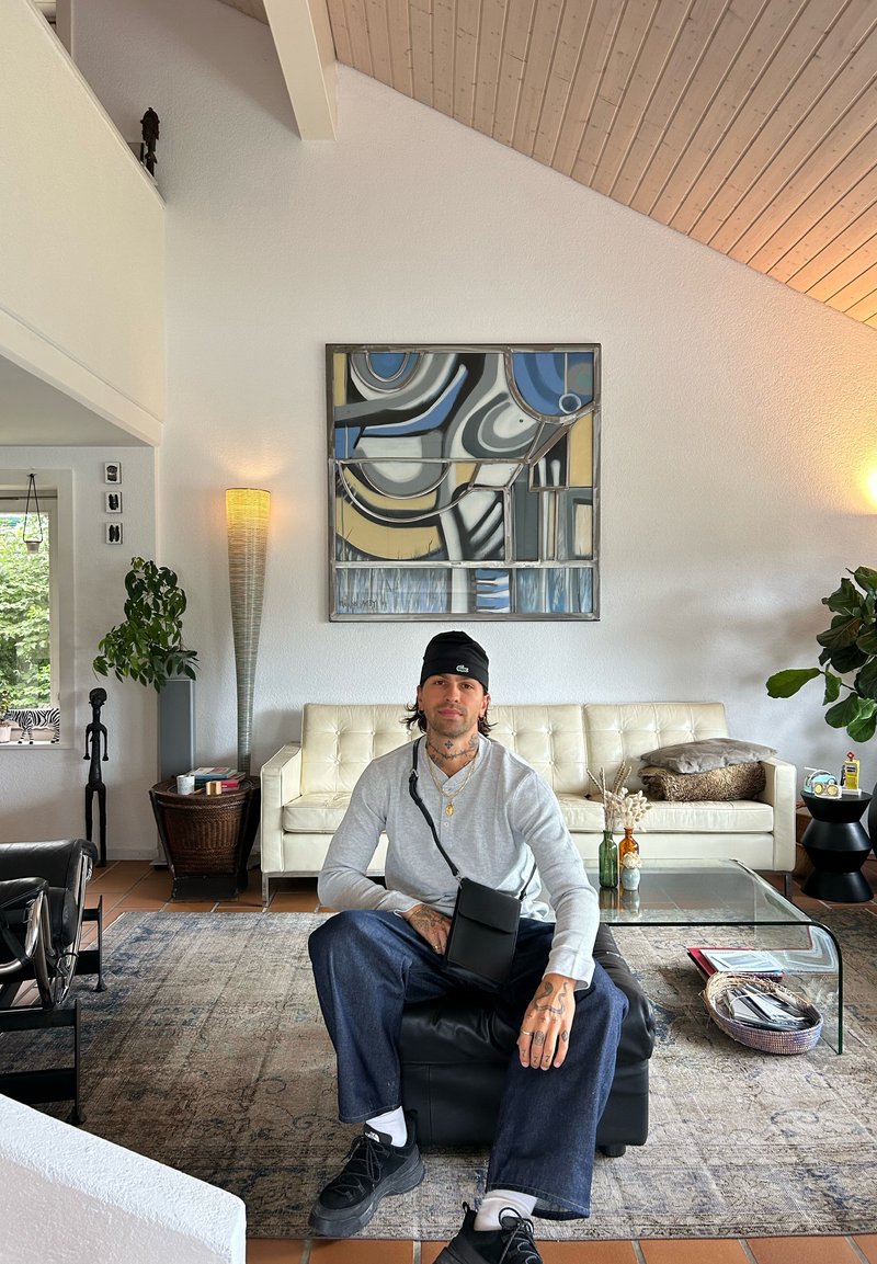 Homem com tatuagens, gorro preto e camisa cinza sentado em um ottomano preto em uma sala de estar moderna com arte abstrata acima do sofá branco.