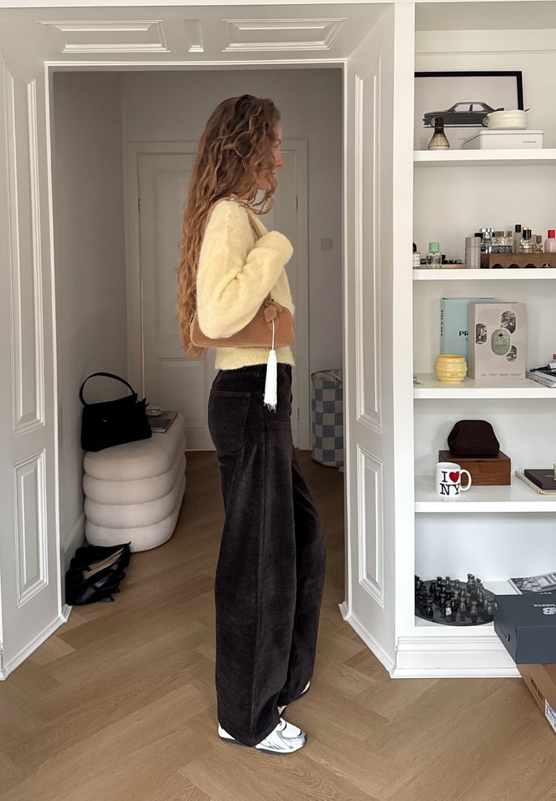 Vrouw in een gele trui en zwarte wijde broek met een bruine schoudertas, staande in een kamer met witte planken en een houten vloer.