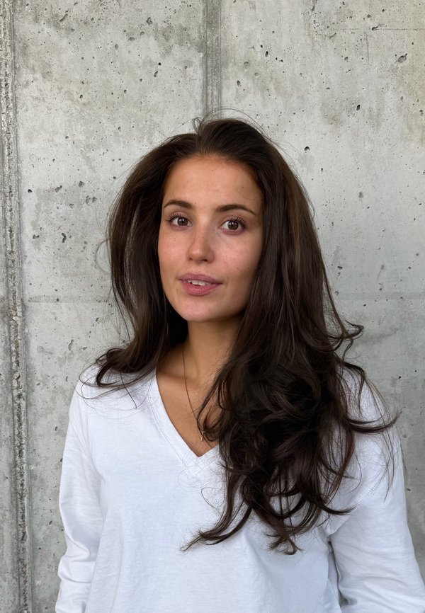 Una mujer de pelo largo y oscuro y una camiseta blanca con cuello de pico está de pie delante de una pared de hormigón gris y mira a la cámara.