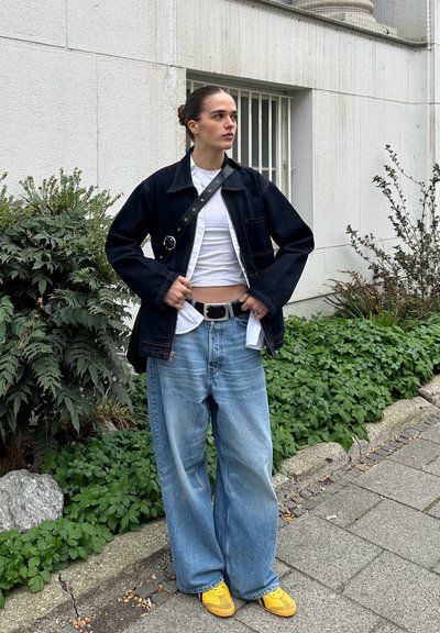 Chaqueta de denim de forma oversized, color navy; camiseta blanca debajo; jeans anchos de color azul claro; zapatos amarillos; fondo urbano con vegetación.