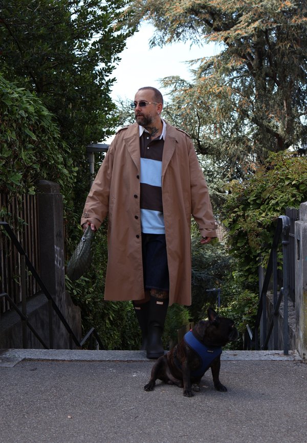 Un hombre con un abrigo beige y botas de goma camina por un sendero del jardín con una correa, mientras delante se sienta un perro de pelaje oscuro con un arnés azul.