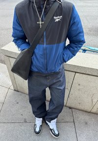 Άτομο που φοράει μπλε και μαύρο μπουφάν Reebok, σκούρο τζιν, άσπρα και μαύρα αθλητικά παπούτσια, ασημένιο κολιέ με σταυρό και μαύρο τσαντάκι ώμου, στέκεται σε πεζοδρόμιο.