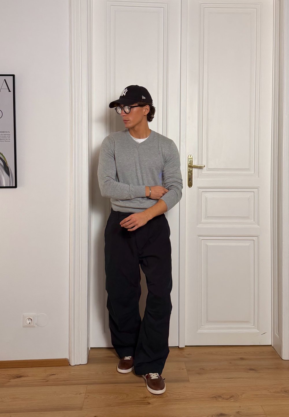 Moški v sivi jopi, črnih hlačah, rjavih supergah in črni kapici stoji pred belimi vrati v minimalistični sobi.