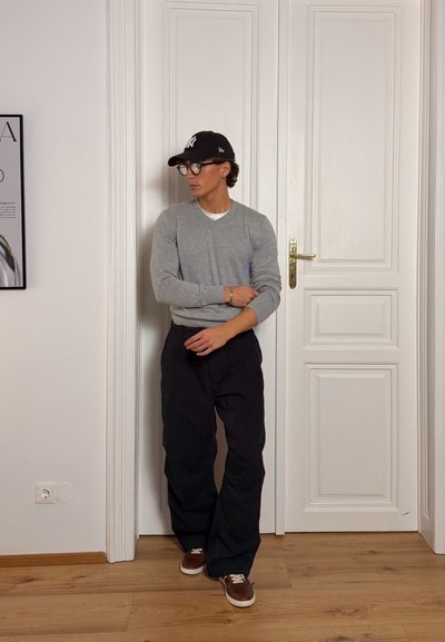 Hombre que lleva un suéter gris, pantalones negros, zapatillas marrones y una gorra negra, de pie frente a una puerta blanca en una habitación minimalista.