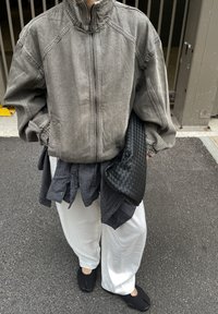 Šedá bunda na zip s volnějším střihem, vrstvená přes černobílou kostkovanou košili, doplněná bílými kalhotami s širokými nohavicemi a černými nazouvacími botami.