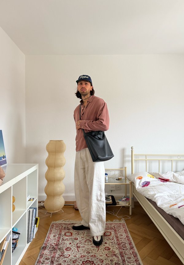 Homme portant une chemise rose, un pantalon blanc, une casquette noire et un sac à bandoulière noir se tient dans une chambre bien rangée avec un lit, une lampe et une étagère.
