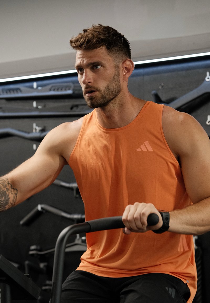 Homme musclé en débardeur orange utilisant du matériel de gym, concentré sur son entraînement, avec des poids et des barres fixés au mur derrière lui.