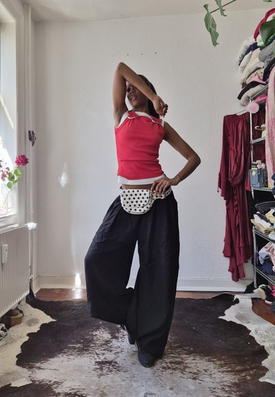 Persona que lleva una camiseta sin mangas roja y pantalones anchos negros, posando con un brazo doblado sobre la cara y el otro en la cadera dentro de una habitación cerca de un perchero.