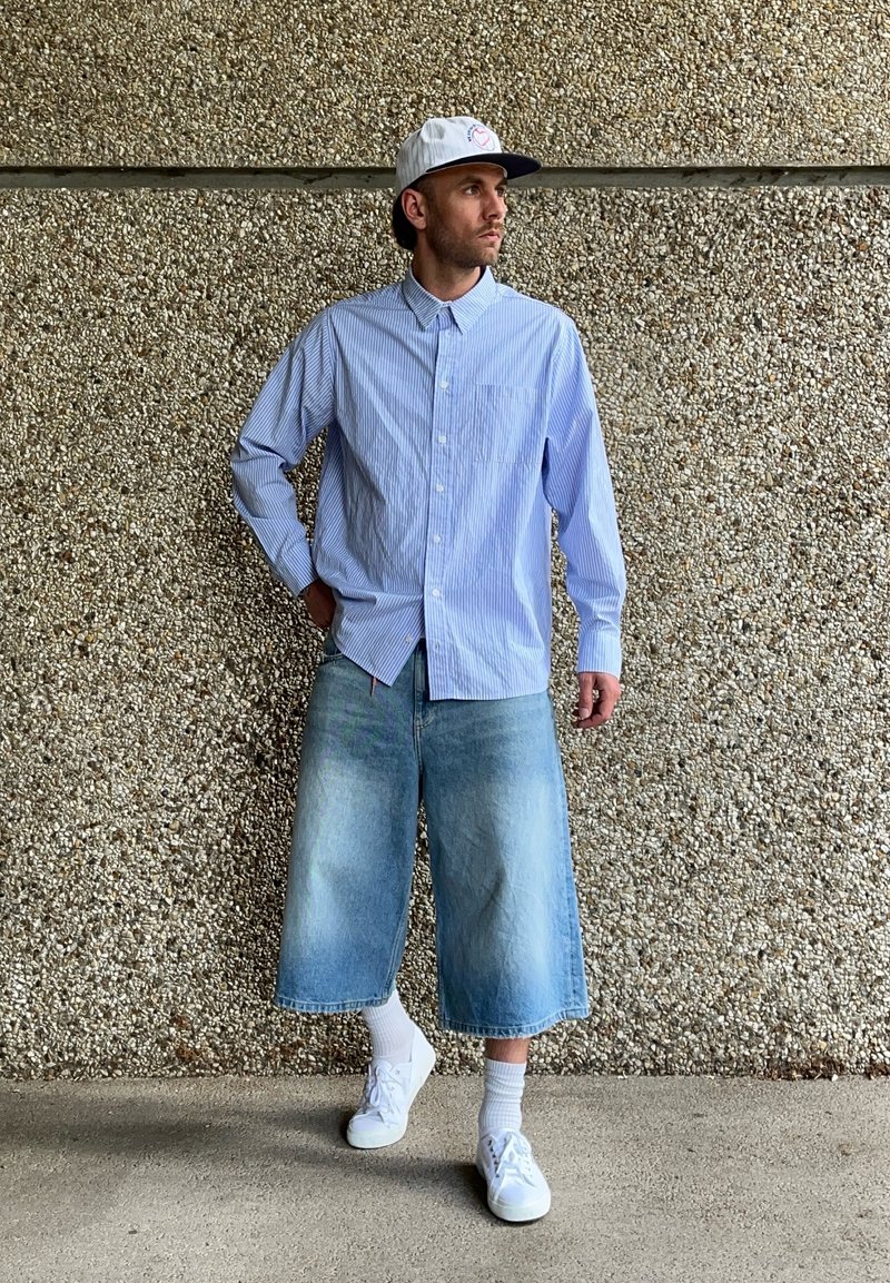 Chemise à rayures bleu clair à manches longues, culottes en denim, baskets blanches et casquette blanche avec logo. En toile de fond, un mur en béton texturé.