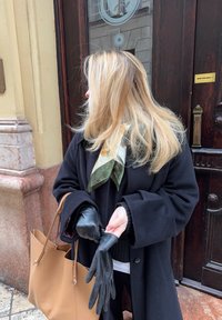 Melnas ādas cimdi ar teksturētu apdari, kurus tur rokās persona, kas valk ā melnu mēteļu un rakstainu zaļo un krēma krāsas šalli, kā arī smilškrāsas somu.