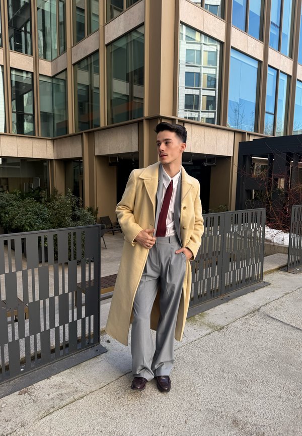 Młody mężczyzna stoi na zewnątrz obok nowoczesnego budynku, ubrany w beżowy długi płaszcz, białą koszulę, czerwony krawat, szare spodnie i ciemne buty.