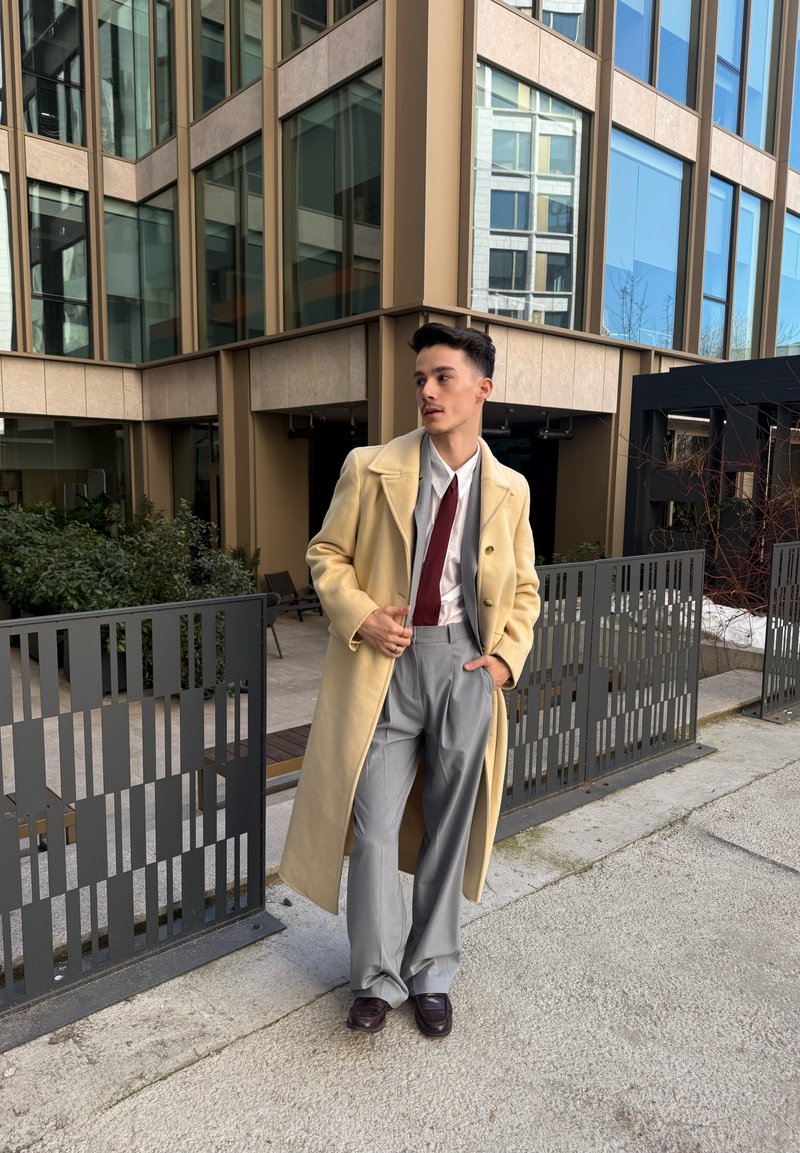 Νέος άνδρας που στέκεται έξω κοντά σε μοντέρνο κτίριο, φορώντας μπεζ μακρύ παλτό, άσπρο πουκάμισο, κόκκινη γραβάτα, γκρι παντελόνια και σκοτεινά παπούτσια.