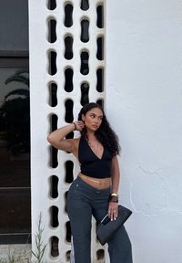 Top negro tipo halter, pantalones deportivos grises con rayas blancas y un bolso de mano negro. Cabello rizado, de pie junto a una pared blanca texturizada con aberturas.