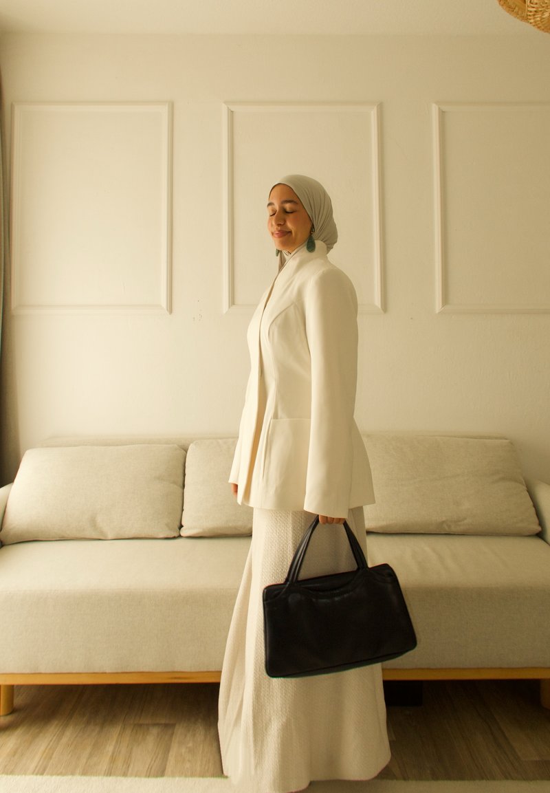 Blazer bianco sopra una gonna chiara e strutturata, con in mano una borsa nera. Lo sfondo presenta un divano neutro e pareti di un colore chiaro uniforme.