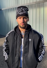 Veste de sport noire avec des bandes blanches, débardeur blanc et bonnet noir avec un motif graphique blanc. Yeux bleus et cheveux courts.