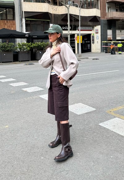 Mujer con chaqueta beige, gorra verde, pantalones cortos oscuros hasta la rodilla y botas marrones de pie en una calle de la ciudad cerca de un paso de peatones.