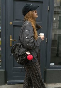 Pelēks džemperis ar pogu akcentiem, melna soma ar atteiktu siksnu, sarkans atslēgu piekariņš un raibu bikses. Kafijas krūze turēta labajā rokā.