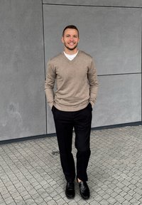 Beige v-neck villapaita, valkoinen aluspaita, mustat housut ja mustat kengät. Seisoo teksturoidulla asfaltilla harmaata seinää vasten.