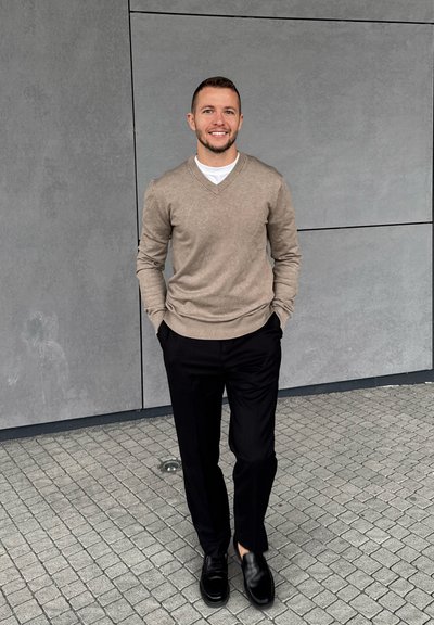 Suéter beige de cuello en V, camiseta blanca debajo, pantalones negros y zapatos negros. De pie sobre un pavimento texturizado contra una pared gris.