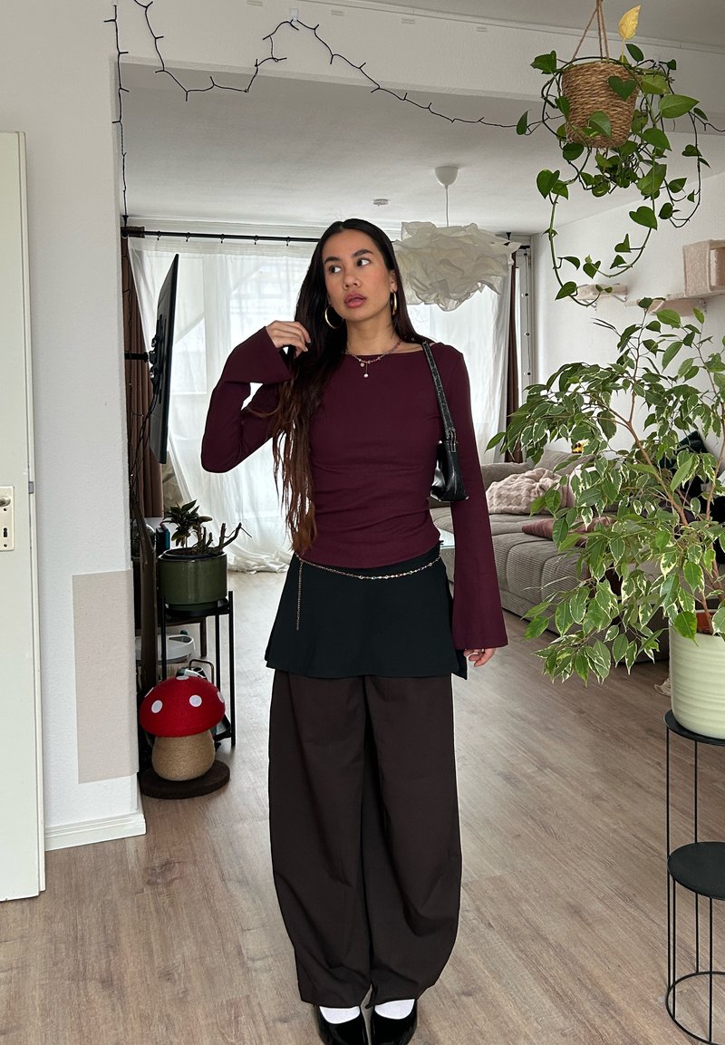 Femme aux longs cheveux foncés portant un haut bordeaux, une jupe noire sur un pantalon large marron, debout dans un salon rempli de plantes avec une lumière naturelle.