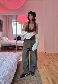 Femme portant une casquette noire et un soutien-gorge de sport avec une veste grise accrochée, debout dans une chambre avec un placard à miroir et une lampe suspendue rose.