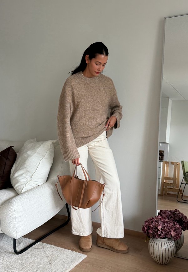 Une femme portant un pull beige, un pantalon blanc et des bottes brunes tient un cabas en cuir brun à côté d’un canapé blanc dans un salon.