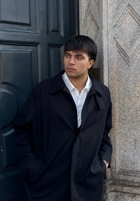 Jeune homme aux cheveux foncés et portant une boucle d'oreille, vêtu d'un manteau noir et d'une chemise blanche, appuyé contre un mur en pierre à côté d'une porte bleu foncé.