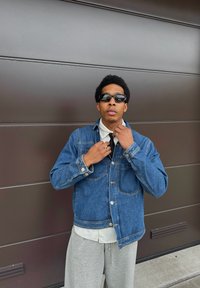 Giacca di jeans blu medio con chiusure a bottoni e tasche sul petto, indossata sopra una camicia bianca e pantaloni grigi, completata da occhiali da sole neri.