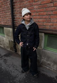 Temno jeans jakna z dvema žepoma na prsih, pod njo siva kapuco, prevelike črne hlače, bela kapa, stoji ob opečnem zidu.