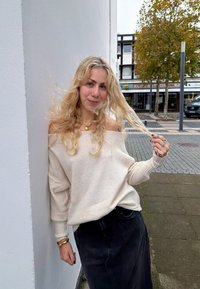 Sweater off-shoulder em bege claro combinado com uma saia preta. Detalhe de um colar com pingente e anéis nos dedos. Fundo urbano descontraído.