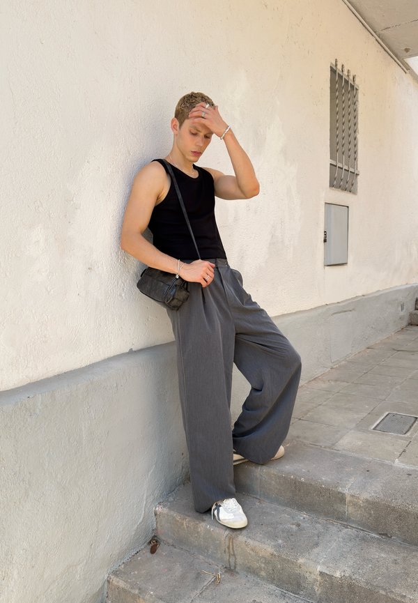 Jeune personne aux cheveux courts appuyée contre un mur blanc sur des escaliers extérieurs, portant un débardeur noir, un pantalon large gris, des baskets blanches et un sac à bandoulière.