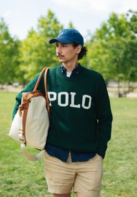 Nuori mies, joka käyttää tummanvihreää 'POLO' -neuletta, sinistä lakkia ja vaaleita shortseja, kantaa suurta beigeä ja ruskeaa kassi, seisoo nurmialueella puiden keskellä.