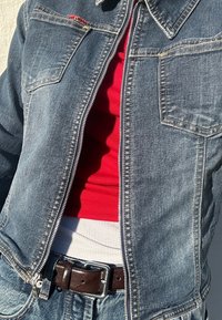 Veste en jean de teinte moyenne avec des accents rouges. Débardeur côtelé rouge et blanc superposé en dessous, complété par une ceinture marron foncé avec une boucle argentée.